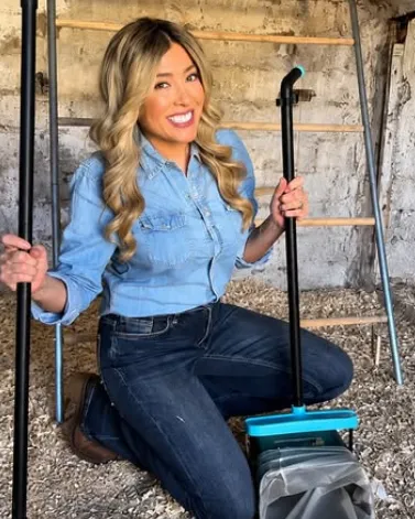 A smiling woman in a denim shirt kneels in a barn with a broom and dustpan, doing sanctuary chores